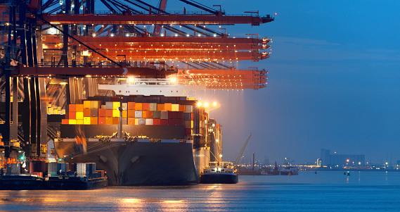 Gantry cranes loading illuminated cargo container ship moored in commercial dock at night, Rotterdam The Netherlands